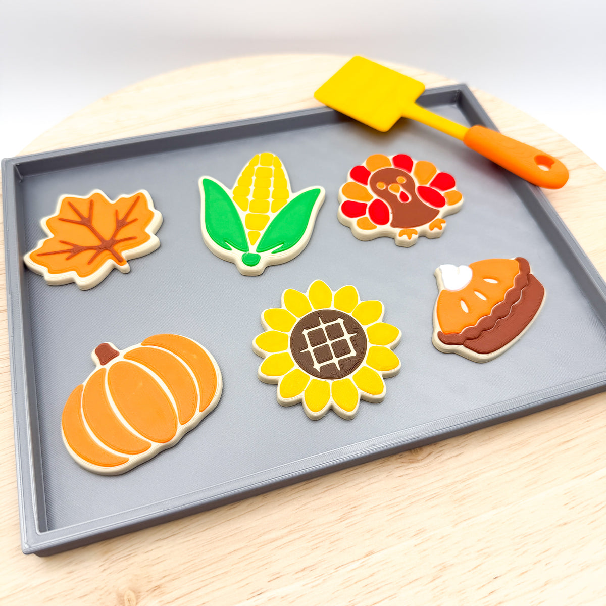Angled view of 6 fall pretend cookies on a gray tray with an orange and yellow spatula resting on the edge