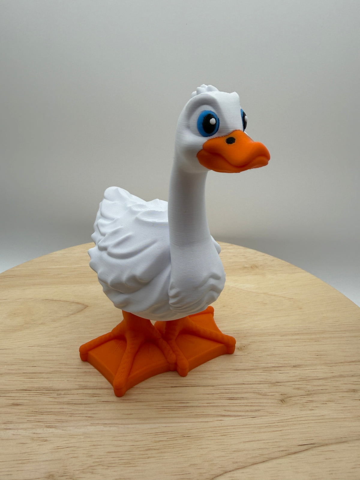 White 3D printed goose with blue eyes and orange beak and feet standing on a wooden surface
